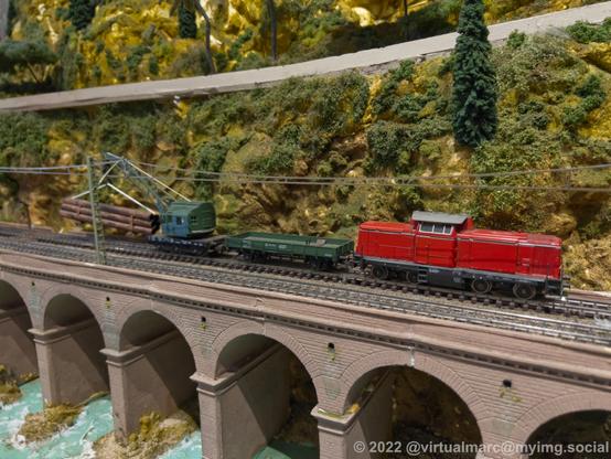 A photo of a bridge of a model railway. A red diesel train carrying a wood crane is on the bridge.