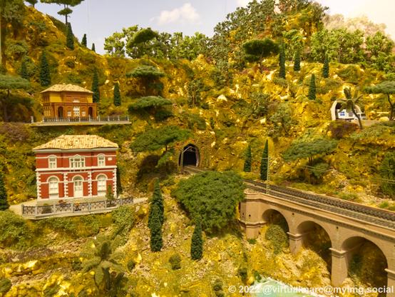 A photo of a hill on a model railway with two houses on it. A railway bridge is going towards the hill and into a tunnel.