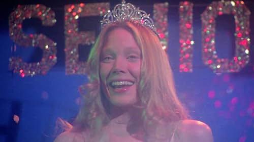 A girl with shoulder length hair smiles widely with a prom queen crown on her head