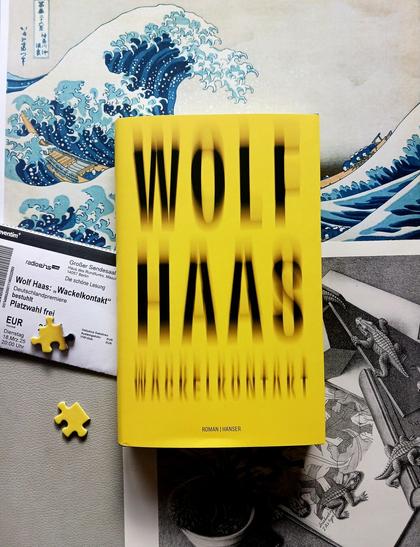 Foto des Buches, auf dessen knallgelbem Cover der Name des Autors und der Titel nur verschwommen zu lesen sind. Das Buch liegt auf zwei berühmten Bildern, zum einen dem farbigen Holzschnitt »Die große Welle vor Kanagawa« von Katsushika Hokusai, zum anderen der Lithografie »Reptilien« von M. C. Escher. Unter das Buch ist der Ausdruck einer Eintrittskarte für die Deutschlandpremiere des Buches im März in Berlin geschoben. Außerdem sind auf dem Foto zwei Puzzleteile zu sehen, ein knallgelbes Randstück und ein schwarz-gelbes Stück mit drei »Ohren«.