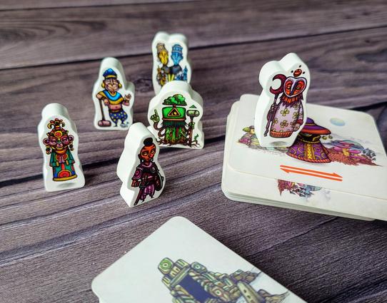 Sechs Meeples bedruckt mit Gestalten aus dem Spiel Faraway stehen auf und neben dem Stapel der quadratischen Karten des Spiels. Im Vordergrund liegt der kleinere Kartenstapel der Heiligtümer
