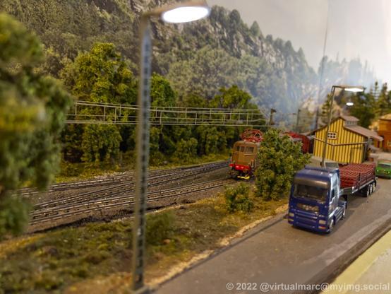 A photo of a model railway with a street next to the rails. There is a freight train approching on the rails and a truck on the streets.