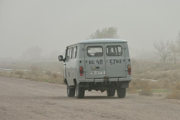 A grey old van travelling through a desert/steppe along a dirty road.