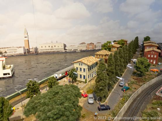 A photo of a coast area of a model railway with some small houses and a train station. There are multiple cars on a narrow street with trees.