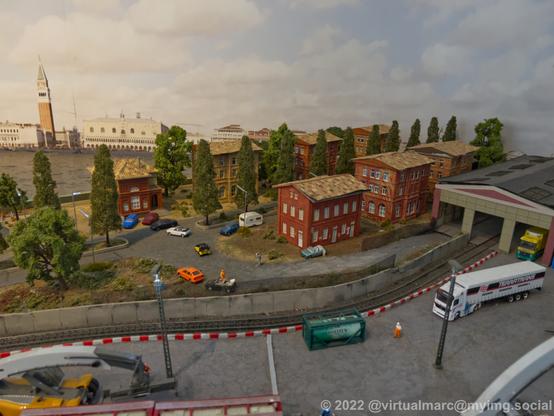 A photo of an industrial complex with a railway connection on a model railway. There are trucks on the complex. Behind are some houses near water.
