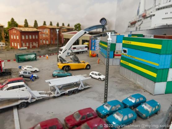 A photo of a model railway at a port with many cars and containers that are loaded and unloaded from ships and trucks.