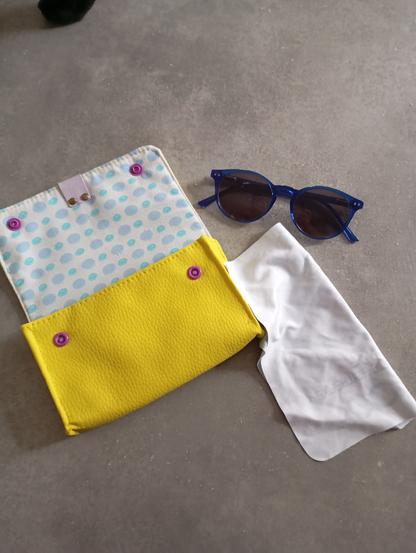 Une pochette jaune citron, doublé d'une tissu blanc à pois bleus et des pressions violette
À côté une paire de lunettes solaires et sa chiffonette.