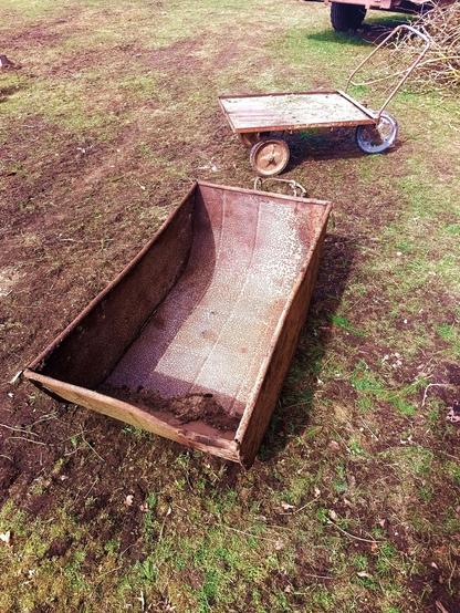 A large metal tub with reinforcement ridges and a piped edge. It's rusty and has some dirt in it and is a little bent at the front. Behind it, a three wheeled steel cart with a flat bed and profiled edge. It has wide handles for pushing around.