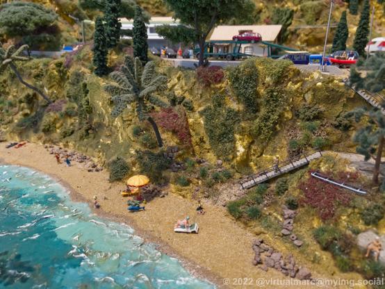 A photo of a beach on a model railway with a street on a hill with cars an trucks on it.