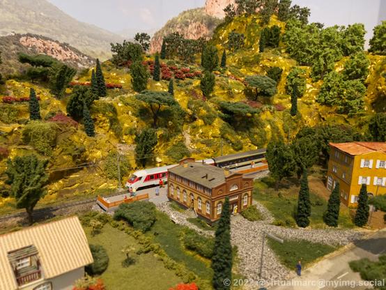 A photo of a small train station of a model railway with a passenger train standing on the paltform. Hills are in the back with trees on it. Some buildings in the front.