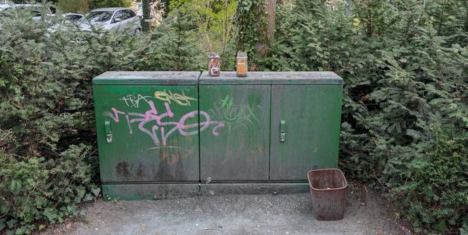 Ein grüner Stromverteiler, auf dem zwei Instantkaffedosen stehen, welche als Aschenbecher genutzt werden. Rechts neben dem Stromkasten steht ein größerer Ascheeimer. Das Ensemble ist von traurig wirkenden Hecken umrahmt.