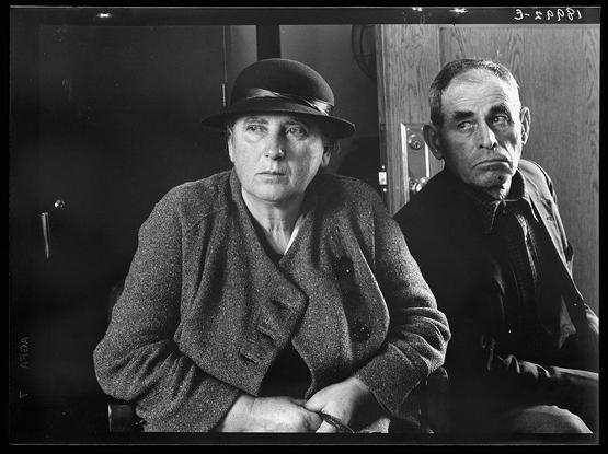 The image is a black and white photograph depicting two individuals, presumably from an earlier era given the style of their clothing. The person on the left appears to be female; she wears glasses, has dark hair pulled back under a broad-brimmed hat, and sports a textured coat that suggests practicality over fashion during challenging times or seasons.

The individual on the right seems male, with short-cropped hair receding from his forehead indicating age. He is dressed in what looks like work clothes - possibly for laborious tasks given their rugged appearance – including a dark jacket and shirt. 

Both individuals are seated closely together against an indistinct background that provides no contextual clues beyond perhaps being inside a building or near one, suggested by the presence of what appears to be a door on the right.

The overall tone is somber; the expressions, though not fully discernible due to resolution constraints, do not suggest happiness. The image captures a moment in time with each subject seemingly absorbed in their own thoughts, conveying an air of solemnity and perhaps economic hardship given historical context often associated such images from early 20th century America.

The photograph is labeled "3-S-4PPI", which could be related to its cataloging or archiving within the collection where it resides. Additional information about this image can indeed be found in a source mentioned, potentiall [...]