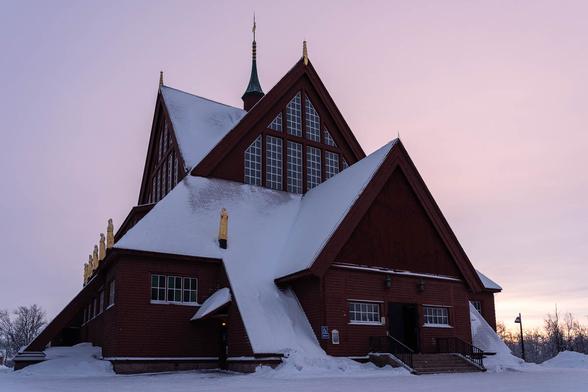 Snow-covered Kiruna Church in Sweden at sunrise, with warm light on its red wooden facade and snow blanketing the roof.