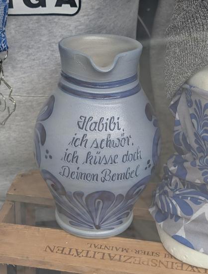 Das Bild zeigt einen blau-grauen #Bembel, der auf einem Holzkarton steht. Der #Bembel hat eine runde Form mit einem breiten Hals und einem flachen Boden. Er ist mit einem dekorativen Muster in Blau und Grau verziert, das sich um den Körper der Vase windet. Auf der Vase ist in deutscher Sprache ein Spruch in geschwungener Schrift eingraviert: "Habibi, ich schwör, ich küsse doch Deinen Bemel." Die Vase steht auf einem Holzkarton, auf dem teilweise der Text "WEINSPEZIALITÄTEN" zu sehen ist. Im Hintergrund ist ein Teil eines grauen Kleidungsstücks und ein blau-weißes Band mit einem Metallhaken zu erkennen.