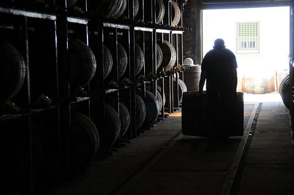 The bonded warehouse at Ardbeg Distillery. The image shows a wide doorway in the upper left of the frame with light pouring into the dark building. On the left are racks holding barrels that are at least three barrels high before disappearing out of the frame. There are tracks on the ground leading to the doorway and a man is rolling a barrel away from the viewer, towards the doorway.