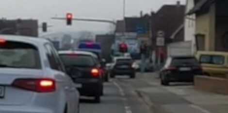 Ein unscharfes Bild einer an einer roten Ampel wartenden Reihe von Autos. Die Straße wird von einem Geh-/Radweg begleitet. Man sieht vor der roten Ampel ein KFZ auf dem Radweg auf die Ampelkreuzung zu fahren, um die Warteschlange zu umgehen und früher abbiegen zu können.