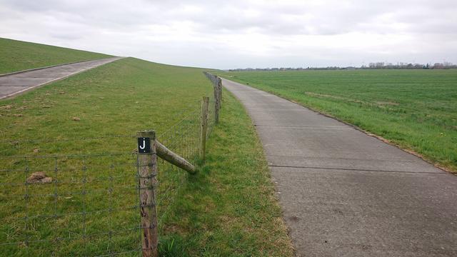 Ein schnurgerader Weg der hinter dem Deich entlang geht. An einem Zaunpfahl ist ein Hinweis auf den Jadeweg.