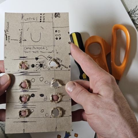 a camera image of a cardboard mock up of an electronic device with the intention of having 8 buttons on the back, and 4 switches or buttons on each side in the thumb posotion and being able to display and or type in braille or utf-8 or other encoded character systems. the cardboard cuttout is 7.25 inches tall and 4 inches wide. there are finger holes cut out at 8 ergonomic intervals simulating button positions on the back of the device. These would be buttons which indicate a 1 or 0 are visually passed through to the person holding it and represent the different character mappings. this could have an wntire screen over it and then be used as an authoring device with eink and potentially haptic feedback, which is one of my goala also long term.