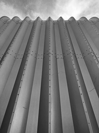 Black and white photo looking up at a corrugated metal wall, the top edge of which curves backward
