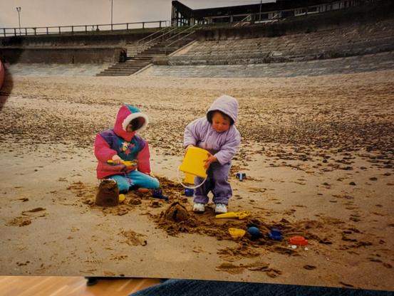 A photo from the 1990s showing 2 girls, aged roughly 5 and 3, making sandcastles on a UK beach on an overcast summer day. Both are bundled up against the cold - wearing long trousers, shoes, and thick parka-style jackets with faux-fur trimming. The post author (Emma) is the girl on the left, barely visible peeking out cheekily from her huge furry hood.