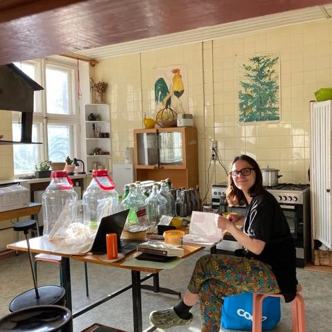 Someone smiling and sitting at at a table in the middle of an old fashion kitchen. On the table: Fermentation containers, unused wine bottles, books, computer.