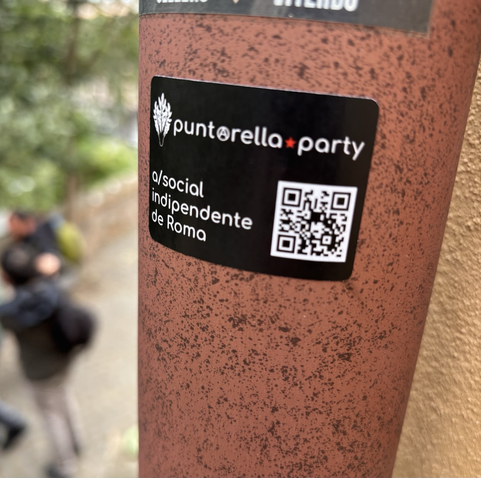 Un adesivo appiccicato su un palo arrugginito. Sull’adesivo il logo di puntarella.party e la scritta “a/social indipendente de Roma”. Di fianco un codice QR che rimanda all’istanza