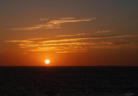 A sunset is reflected in long, thin clouds, above a calm sea.