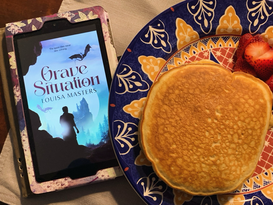 A kindle showing the cover of grave situation next to a plate of pancakes and strawberries