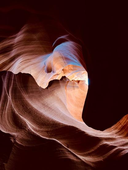 A view of the smooth, swirling sandstone walls inside Antelope Canyon in Arizona. The rock formations display rich hues of orange, brown, and mauve. Sunlight filters in from above and casts an ethereal glow, highlighting soft curves and striations. A faint blue tint appears where the light hits the rock at certain angles, creating a surreal, painterly effect.
