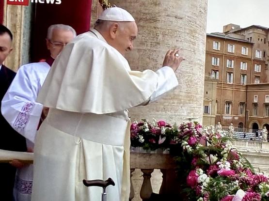 The image shows the pope in white robes, greeting the crowd while standing next to a floral arrangement. A cane is placed beside them, and there are other individuals in clerical attire nearby. The setting appears to be outdoors in a historical location.