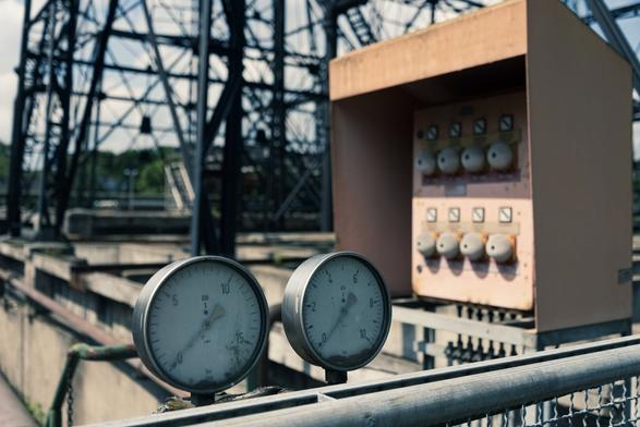 Zwei Manometer werden im Vordergrund angezeigt, mit Messwerten auf einer Skala. Im Hintergrund befindet sich ein Bedienfeld mit mehreren Tasten und Anzeigen, die vor einer Industriestruktur stehen.