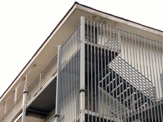 Vista oblicua de un edificio haciendo esquina. Su suave color blanco hueso contrasta con las barandillas grises de sus balcones y las escaleras de emergencia. Éstas, en el lado derecho de la imagen, trazan líneas en horizontal y vertical que se cruzan.