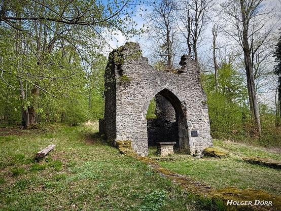 Rückansicht der Ruine St. Valentin. Der Spitzbogen der ehemaligen Altarseite öffnet sich nach außen in den grünen Wald. Ein schlichter Altar steht im Inneren. Links daneben eine einfache Holzbank, rechts und links ist die Ruine von Moos überwuchert. Die Szene wirkt ruhig und verwunschen.
