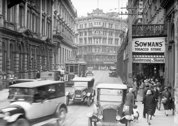Black-and-white photo: Lambton Quay, looking towards Willis Street. 1920s. Description: Lambton Quay, looking towards the New Zealand Railways Booking Office at the intersection of Featherston and Willis Streets. There are cars and a tram travelling down Lambton Quay towards the photographer, and a sign advertising the premises of Sowman’s Tobacco Store is on the right side of the street. Photographer, Sydney Charles Smith. Citation: Smith, Sydney Charles, 1888-1972: Photographs of New Zealand. Ref: 1/2-047590-G. Alexander Turnbull Library, Wellington, New Zealand. https://natlib.govt.nz/records/23136054