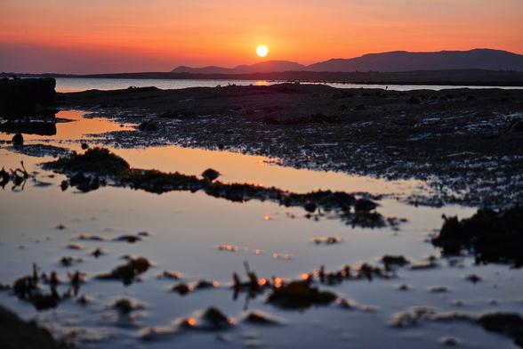 A wide shot of a tranquil landscape at sunset, featuring still water reflecting the vibrant orange and pink sky and the silhouetted distant hills. The foreground shows dark, textured ground with small pools of water mirroring the sunset.