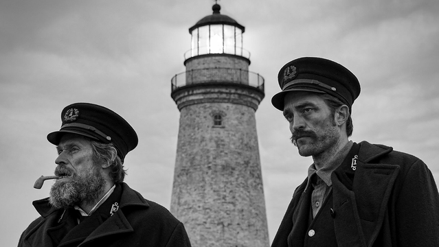 A black and white still from the film "The Lighthouse" shows two men in period clothing standing outdoors with a tall lighthouse behind them. On the left, a man with a weathered face, a long, untamed beard, and a dark cap with an emblem smokes a pipe. He is looking off to the left. On the right, a younger man with a mustache and a serious expression wears a similar dark cap and coat. He is looking slightly to the right. The lighthouse is a cylindrical stone structure with a bright light emanating from the top. The sky is overcast and dramatic.