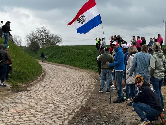 Toeschouwers met een Nederlandse vlag in de berm naast een landweg met kasseien.