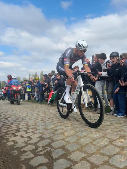 Wielrenner (Mathieu van der Poel) rijdt solo over een kasseienweg gevolgd door een motor. Aan de kant van de weg juichende supporters.