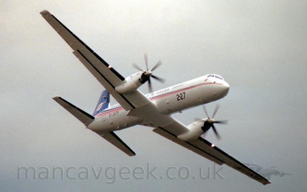 Side view of a white, twin propellor-engined flying from left to right at a low altitude, it's wings banked to the left, with flaps lowered slightly behind the wings, and it's undercarriage stowed away.
A bright light is shining forwards about halfway along each wing.
The fuselage is mostly white, with a thin blue, red, and orange stripe running along the body from the nose, gradually getting wider as it moves rearwards, until the blue sweeps up to fill the tail.
The number "227" is on the lower forward fuselage in large black print, with the much smaller registration "G-JATP" on the lower rear fuselage.
Grey cloudy sky fills the rest of the frame.