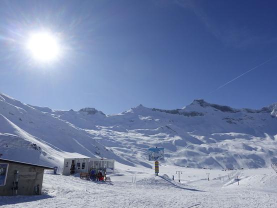 The ski resort of Engstligen Alp. Some lifts and pists in the background. A blue sky and a bright sun on the sky. On the left a grey hut with a pair of white HEAD skis leaning on its wall. A little bit to the right a white pavillon of the skiing school. Some skis and sleds leaning on a wooden ski holder. In the center a plan of the lift and pists with red yellow and blue eplaining signs. Just the very top of the surrounding mountains is not snow white but rocky.