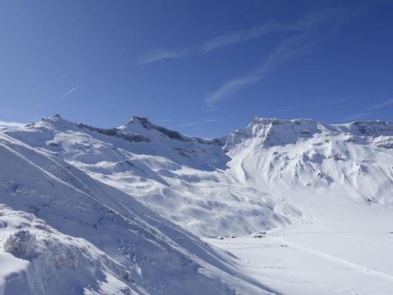Looking down to the ski ressort from a little bit further up the mountain. In the foreground on the left snow-frozen grass and a thin layer of fresh snow. In the background the snowy mountains. In the center the summit of the "Stäghore" as far as I can tell.