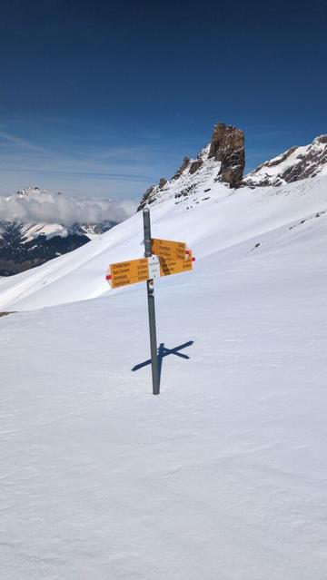 In the foreground a swiss hiking sing at Engstligengrat 2615m. Red-White to the left "Chindbettipass", "Rote Chumme", "Gemmipass", "Engstligenalp". Red-White to the right "Engstligenalp", "Inner Üschene", "Kandersteg", and in direction of the photographer red-white to "Täli", "Schwarenbach", "Inner Üschene". In the background at the center the rocky summit of the "Tschigellochtighore". On the left down in the green valley far away some houses of Adelboden. The mountains behind Adelboden are clouded under a blue sky.