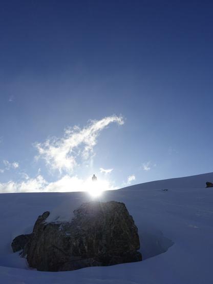 A rock in between the snow. Above the sun and above the sun the upper part of a person. The lower part is overshown by the sun. Some thin cl