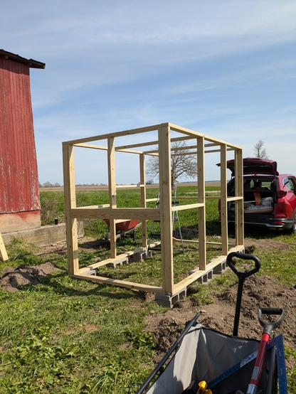 A photo of a bare wooden frame amounting to four walls. The structure is outside on green grass, a bit of a red barn is visible to the side, a utility cart is in the foreground, and a suv with an open trunk is visible in the background. The sky is a beautiful blue.