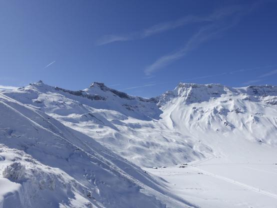 Looking down to the ski ressort from a little bit further up the mountain. In the foreground on the left snow-frozen grass and a thin layer of fresh snow. In the background the snowy mountains. In the center the summit of the "Stäghore" as far as I can tell.