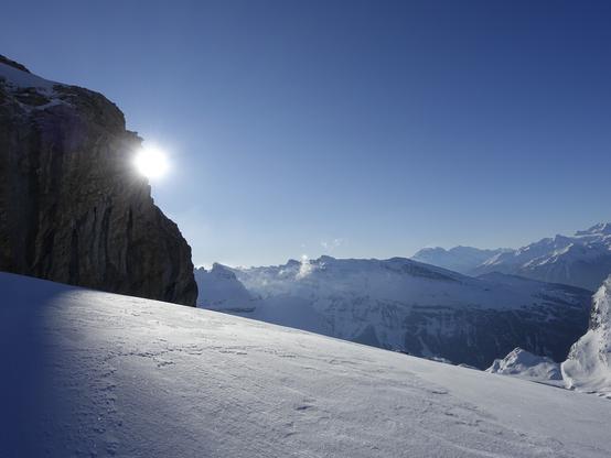 The sun behind a rocky cliff illuminates the drift snow in the foreground. Tiny snow structures look a little bit like the distant moon surface.
