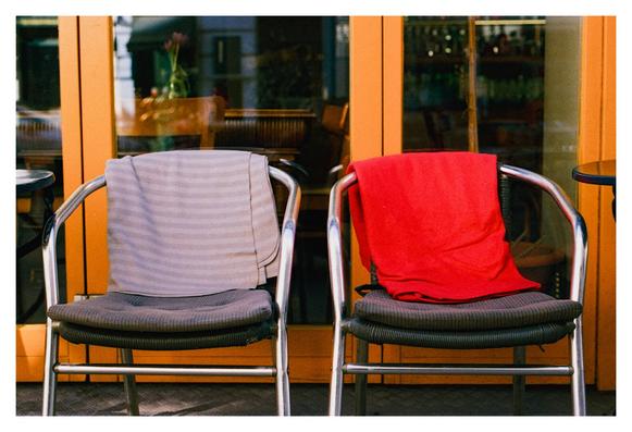 Color photograph of two chairs standing in front of the glass door of a restaurant. A gray striped blanket lies on the left chair. A red blanket is draped on the right-hand chair. The door frame is painted yellow. The wooden furniture of the restaurant interior and a flower can be seen through the glass.