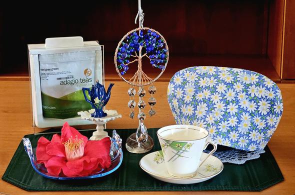 Tea tableau centering around a green and blue jewel beaded Tree of Life suspended on a display stand. The tea cozy is field of daisies, the vintage cup and saucer have a daisy design, and there is a large dark red Japanese Camellia blossom on an aquarium blue Depression Glass dish. There is a miniature dark blue teapot on a scalloped pedestal, and the tea is Earl Grey Green tea from Adagio.