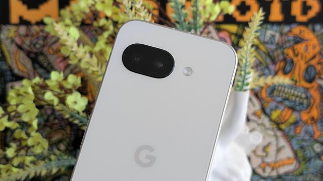 Photo of the back panel of a white Google Pixel 9a (upper 2/3rd, including the black glass camera pill and grey "G" logo) at an angle, with yellow flowers and orange poster out of focus in the background