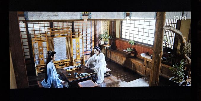 Tsnghua and Yuanzhong drinking wine in a room with framed silk windows. The floor is brown, whereas the low cupboards along the wall is in a lighter brown shade - both things have been worn smooth through ages of use. The light from the windows is falling on a calligraphy scroll hung behind a houseplant, and a yellow and white silk screen on feet pushed to the side.